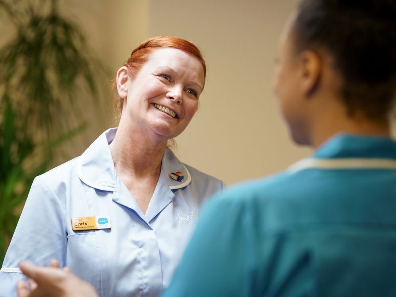 A nurse is smiling at another nurse, whose back is in the foreground of the image.