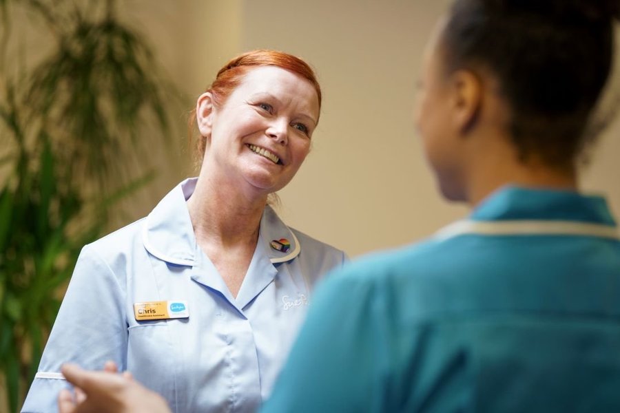 A nurse is smiling at another nurse, whose back is in the foreground of the image.