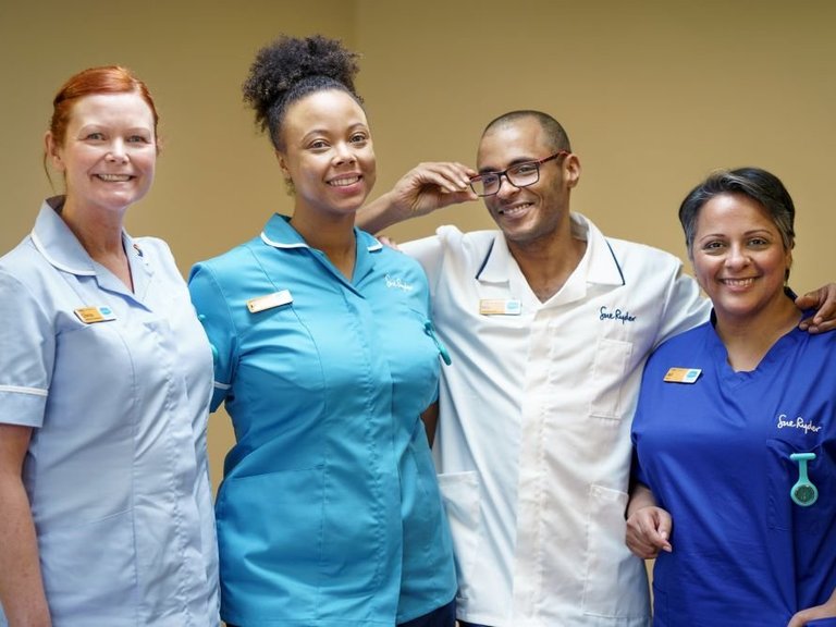 Four healthcare professionals are stood together smiling at the camera. They are a diverse group and each has a different Sue Ryder uniform on demonstrating their role.