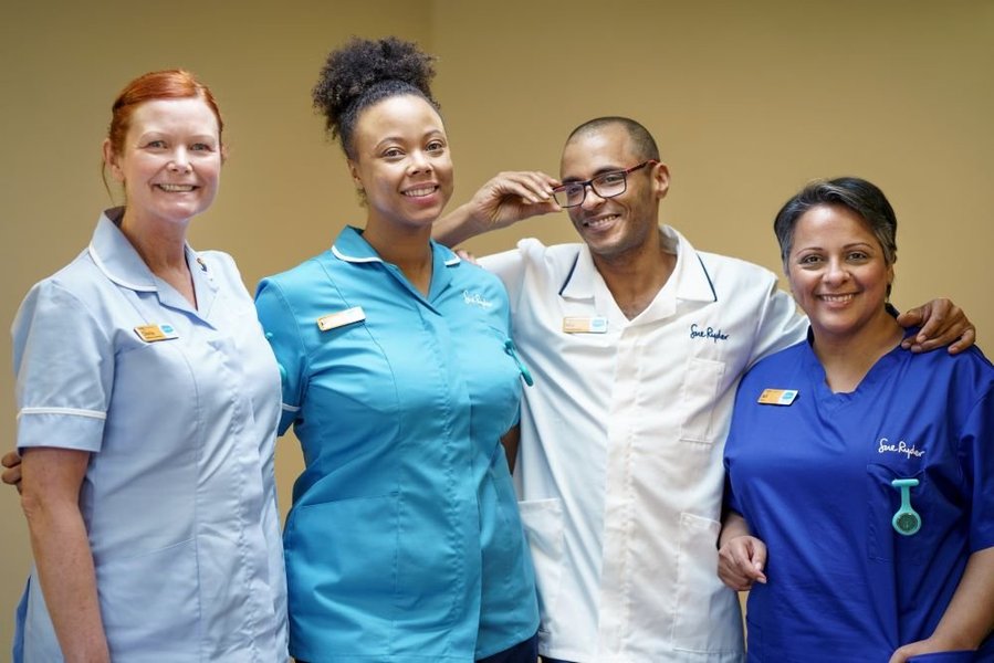 Four healthcare professionals are stood together smiling at the camera. They are a diverse group and each has a different Sue Ryder uniform on demonstrating their role.