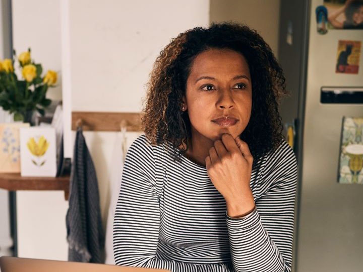 A woman is sat a table looking into the middle distance. In front of her on the table is a laptop and a mug.