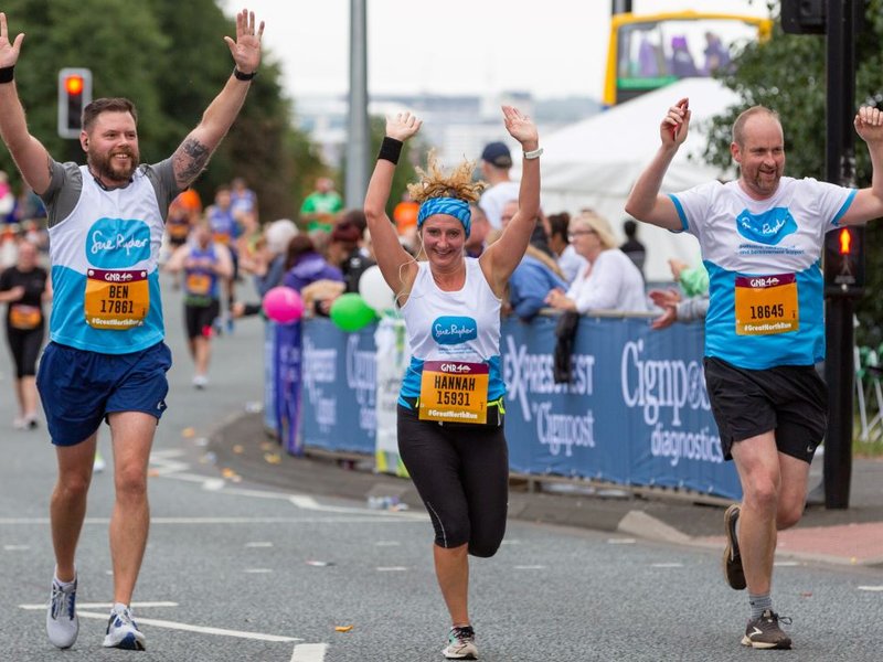 Three runners in Sue Ryder vests are running towards the camera with their arms in the air