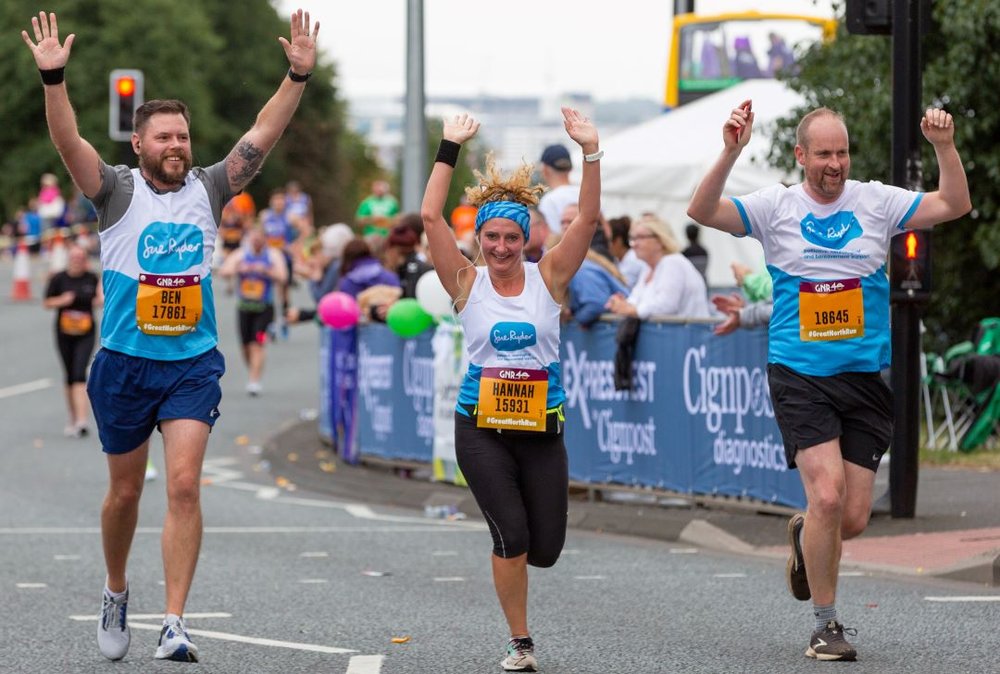Three runners in Sue Ryder vests are running towards the camera with their arms in the air