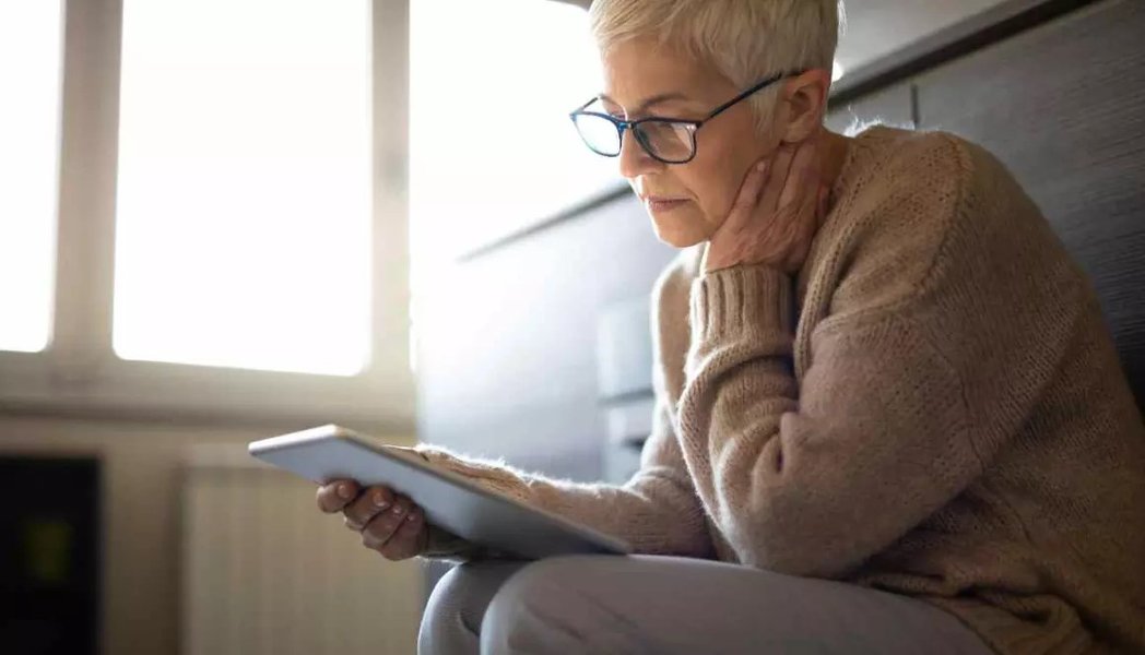 A woman with short grey-white hair and glasses is looking down at a tablet