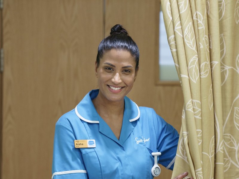 Image of a female Sue Ryder Nurse in blue uniform standing next to a curtain around a bed on a hospice ward