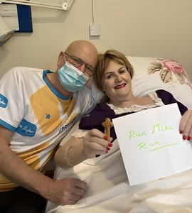 Man sits next to his wife who is lying in a hospice bed. Man is wearing a mask but smiling and woman is smiling, holding a wooden cross in one hand and a paper note in the other reading 'Run Mike Run'