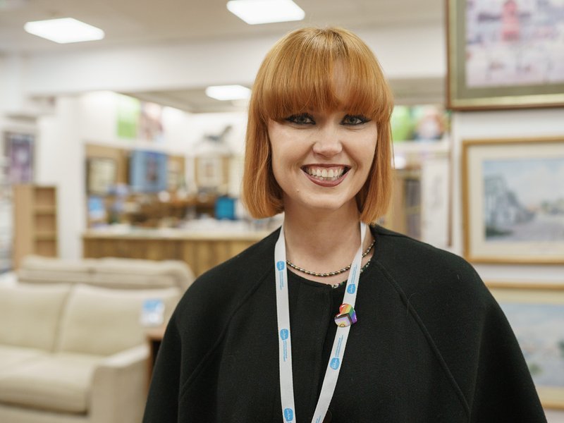 A retail volunteer is smiling at the camera. In the background is a Sue Ryder shop with donated furniture and paintings.