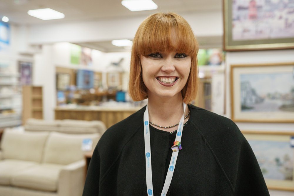 A retail volunteer is smiling at the camera. In the background is a Sue Ryder shop with donated furniture and paintings.