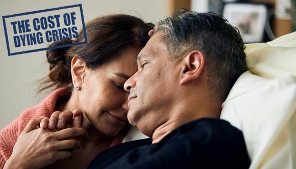 A woman holds her husband's hand as he lies in a hospice bed and a stamp with text saying 'The Cost of Dying Crisis' in the top left