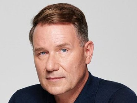 Richard looks directly at the camera with a half-smile, he's against a grey background.