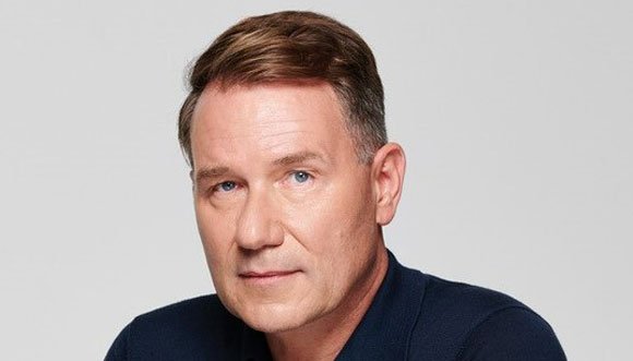 Richard looks directly at the camera with a half-smile, he's against a grey background.