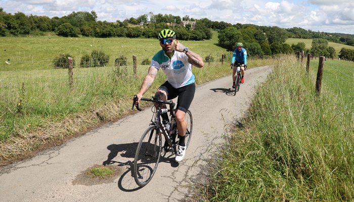 Two riders in their Sue Ryder jerseys, cycling along a country road with fields all around them