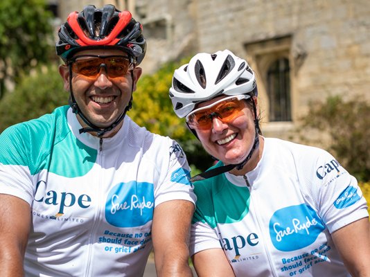 Two cyclists in their helmets and Sue Ryder cycling jerseys, smiling and hugging
