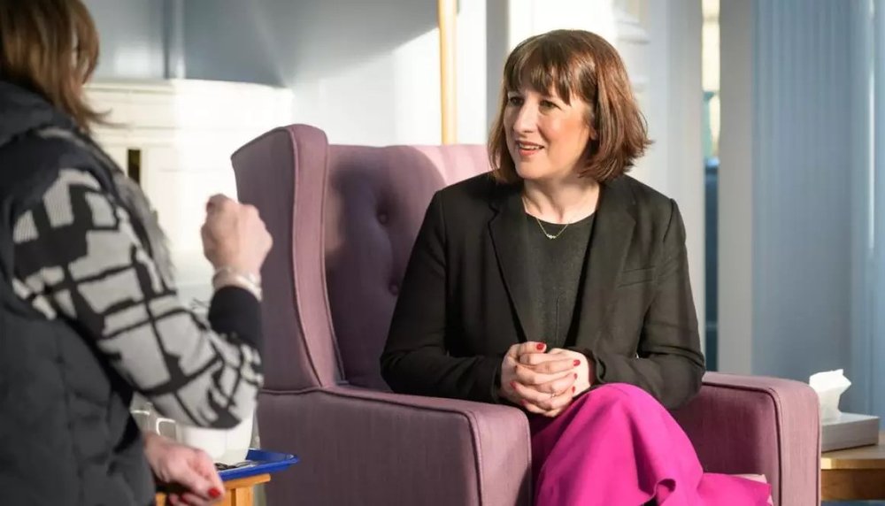 Rachel Reeves MP sits on a pink sofa.  She has mid length brunette hair.  She is speaking to Jane, who we can only see from the back.