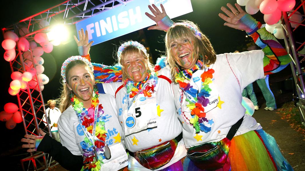 Three smiling Starlight Hike walkers in rainbow clothing and face paint, at the finish line with their medals