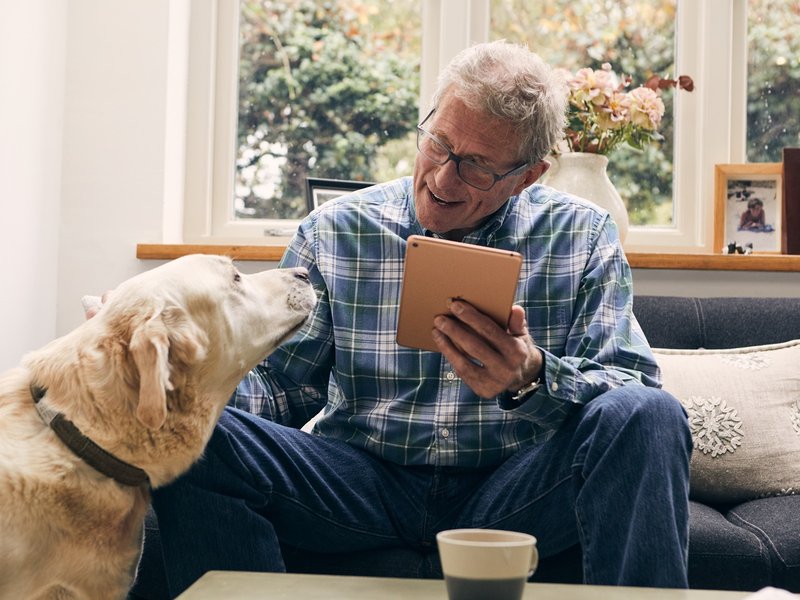 An older man is sat on a living room sofa holding an ipad and with a mug on the table in front of him. He's looking at a yellow dog at his side and has his hand up to pet the dog.