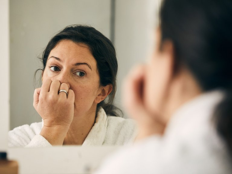 A woman looks into a mirror with her hand up to her mouth.