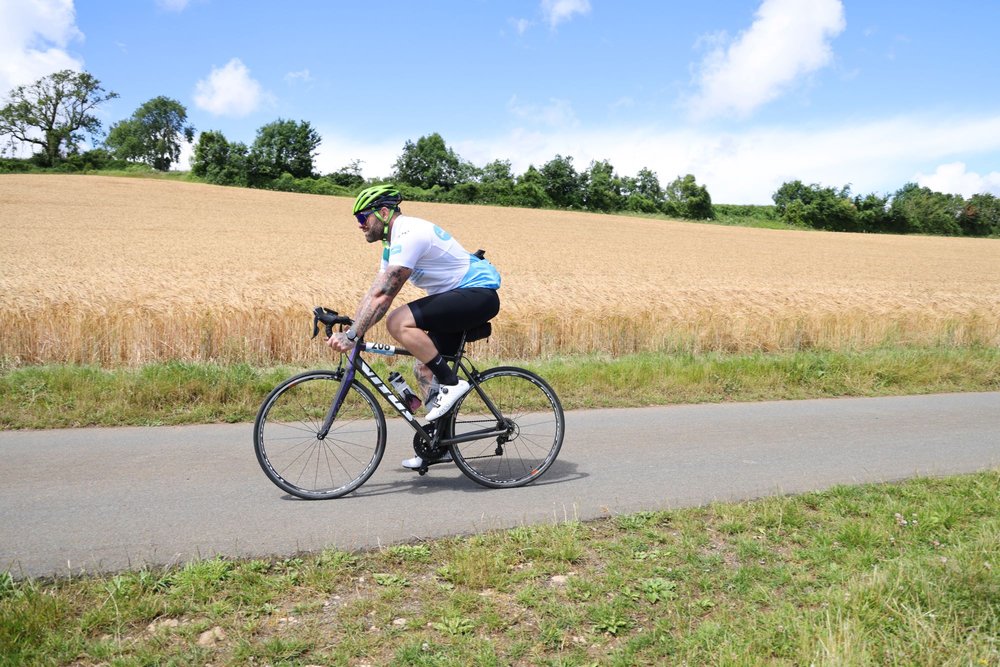 A Sue Ryder cyclist going along a country lane, with golden fields behind them