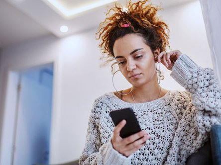 A woman in a grey jumper is looking at a black phone in her hand.