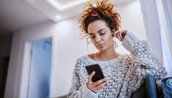 A woman in a grey jumper is looking at a black phone in her hand.