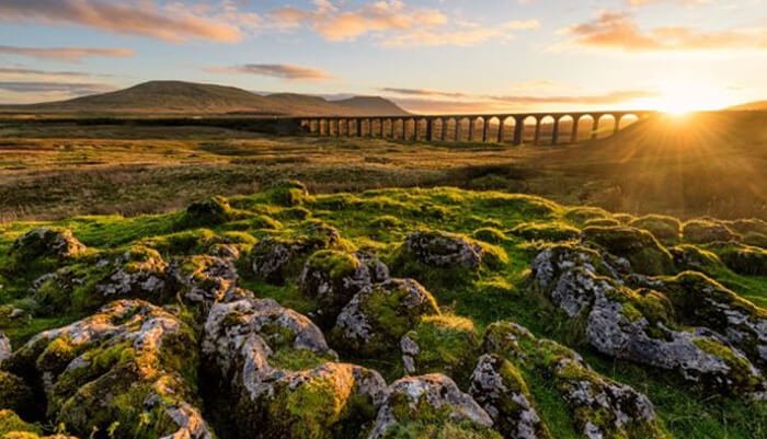 Mossy rocks in the foreground, with a large stone viaduct in the background and the setting sun