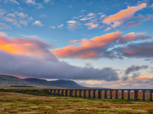 Grassy fields in the foreground, with a large viaduct running through the hills as the sun sets