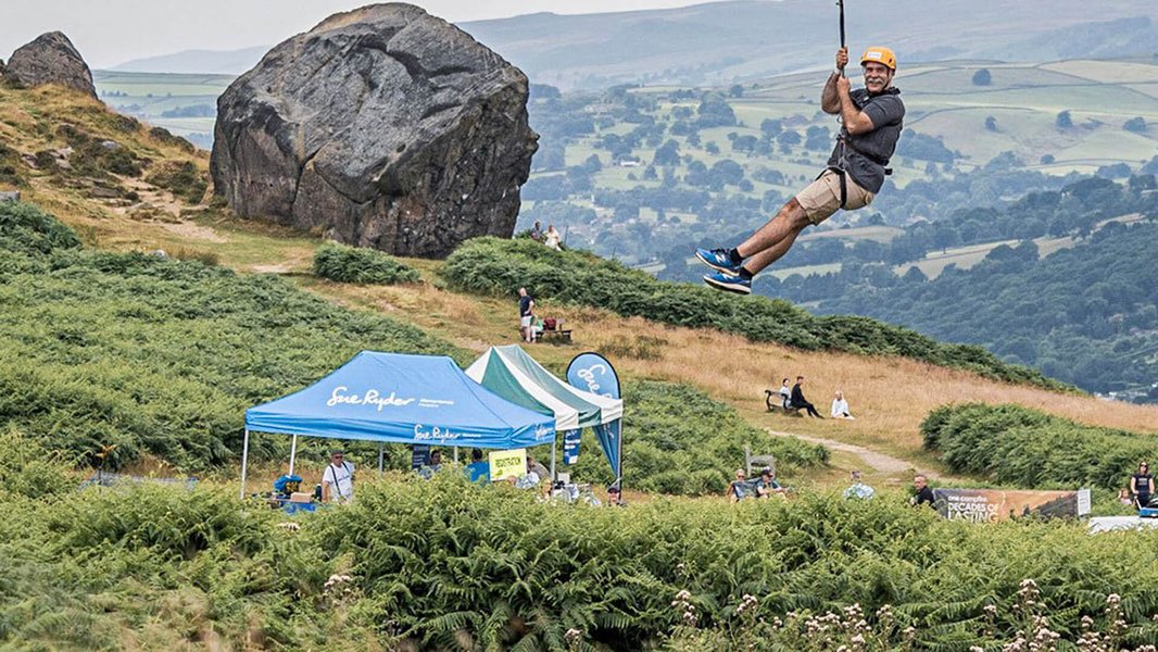 A person zip wiring down from The Cow rocks, with a Sue Ryder tent and hills in the background