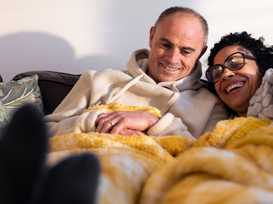 A smiling man and woman snuggle under a blanket on a sofa.