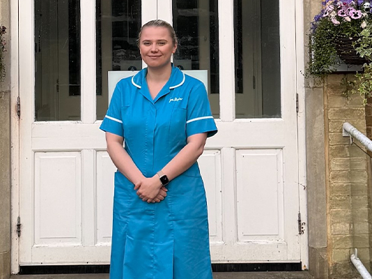 Ellen stands, dressed in her Sue Ryder uniform, in front of white double doors at Sue Ryder St John's Hopsice.