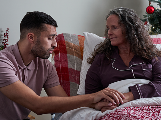 A woman holds in bed clasps hand of younger man next to her. Christmas tree in background.