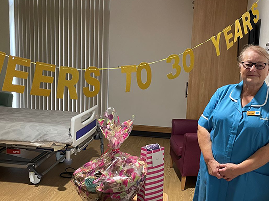 Frances leans against a wall, next to a gold banner tht says 'cheers to 30 years'