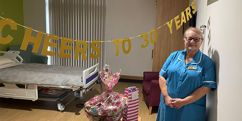 Frances leans against a wall, next to a gold banner tht says 'cheers to 30 years'