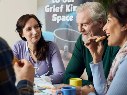 Photo captures 4 people meeting at a Sue Ryder Grief Kind Space, sat around a table whilst chatting, drinking hot drinks and eating biscuits. One female volunteer, one female guest and two male guests