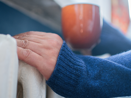 Close up of a person wearing a blue cosey jumper touching a radiator and holding a steaming mug.