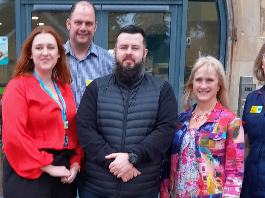 Group of people, including a nurse in Sue Ryder uniform, stand in front of a hopsice entrance.