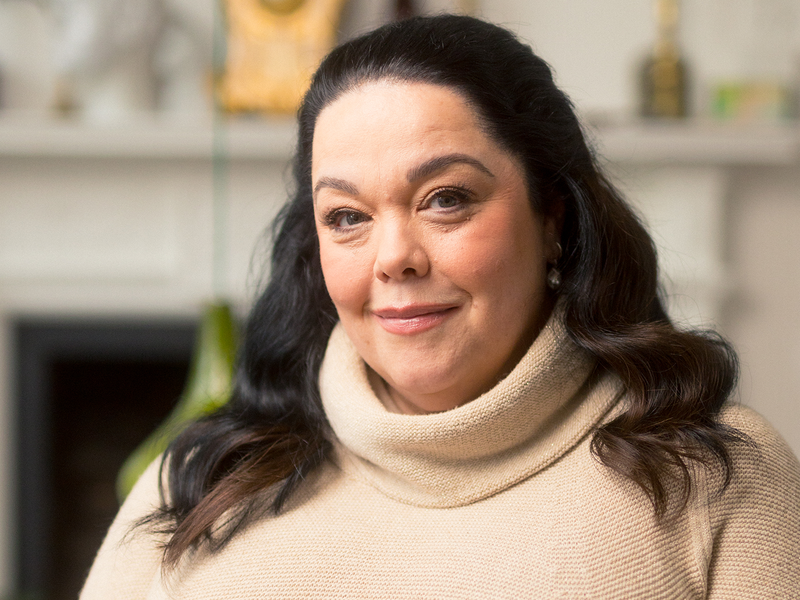 Lisa Riley smiles into camera in front of soft-focus fireplace