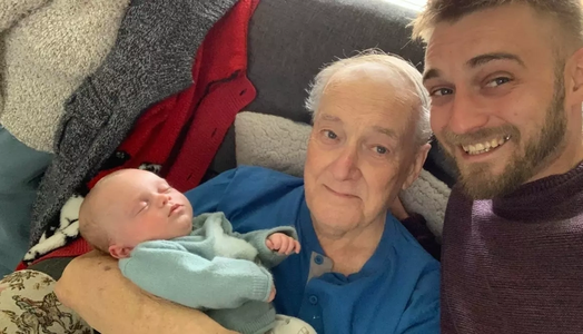 Son stands next to his father who is sitting on a sofa and holding a sleeping baby, his grandson. Both father and son are looking at the camera and smiling.