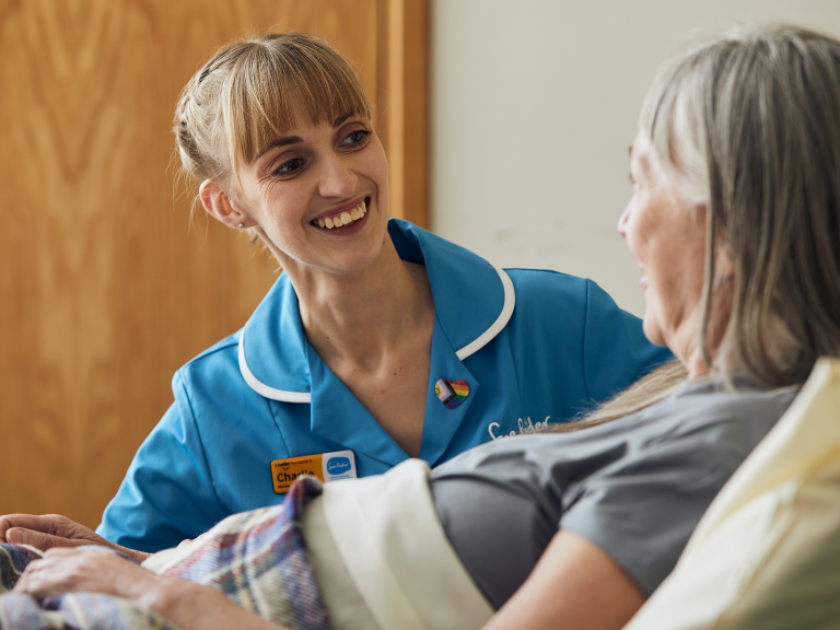 A woman laying in bed while a nurse in blue Sue Ryder uniform smiles broadly at her.
