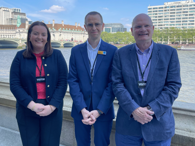Three people pose smiling in front of the River Thames near parliament