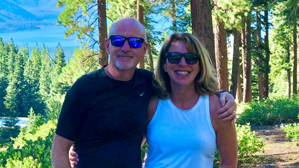 Phil and Anna stand in a rich green forest with water in background
