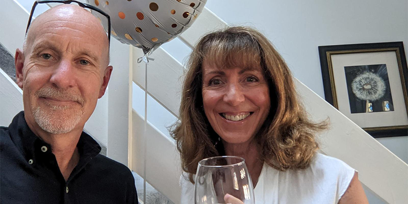 Phil and Anna smile into a camera holding a glass of wine and anniversary balloon