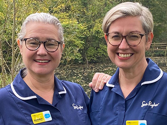 Rachel and Annabel, wearing blue Sue Ryder nurse uniforms, smile into the camera. Annabel rests her hand on Rachel's shoulder.