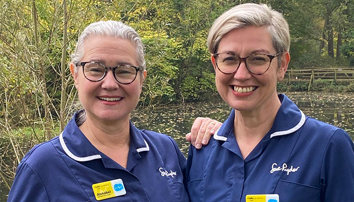 Rachel and Annabel, wearing blue Sue Ryder nurse uniforms, smile into the camera. Annabel rests her hand on Rachel's shoulder.