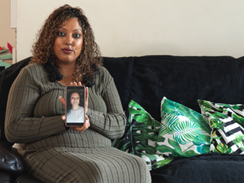 Shireen sits on sofa, holding phone with a photo of her mum on it.