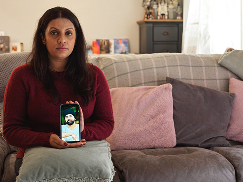 Sunita sits on a sofa holding up her phone which shows a picture of Tony