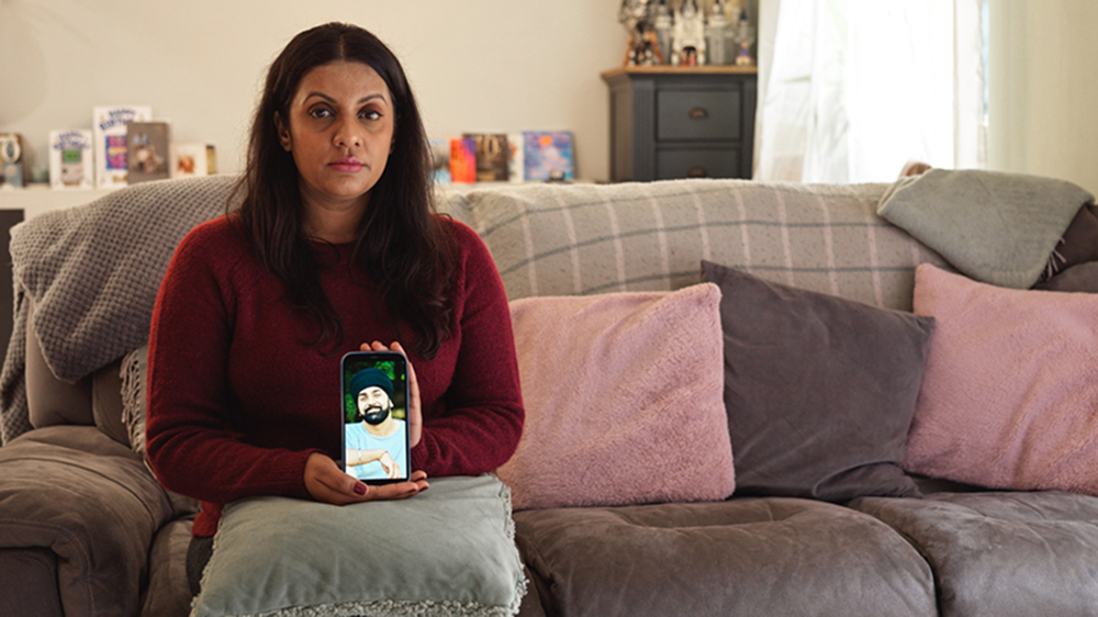 Sunita sits on a sofa holding up her phone which shows a picture of Tony